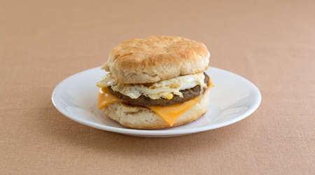 A sausage egg and cheese biscuit breakfast sandwich on a white plate upon a tan table cloth.の写真素材