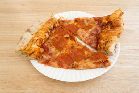 Two slices of freshly baked pepperoni pizza on a white paper plate atop a wood table top illuminated with natural light.の写真素材