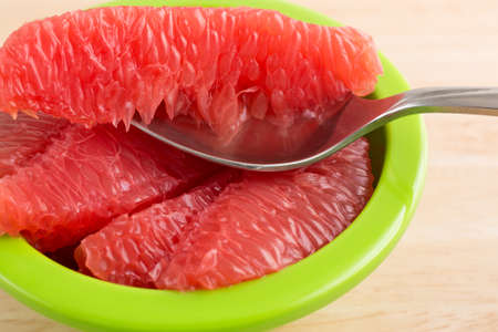 Close view of sections of red grapefruit in a green bowl with a spoon on a wood table top.の写真素材