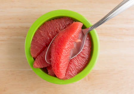 Top view of sections of red grapefruit in a green bowl with a spoon on a wood table top.の写真素材