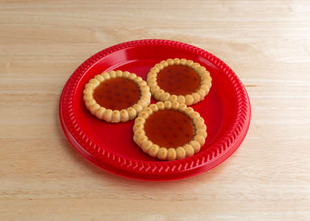 Three strawberry filled tarts on a red plate atop a wood table top illuminated with natural light.の写真素材