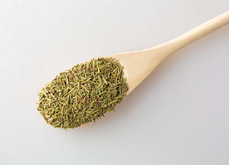 Top view of a portion of dried and chopped rosemary leaves in a wood spoon on a white cutting board...