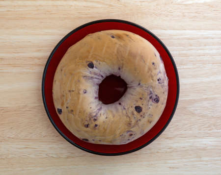 Top view of a freshly baked blueberry bagel on a small red dish atop a wood table top.の写真素材