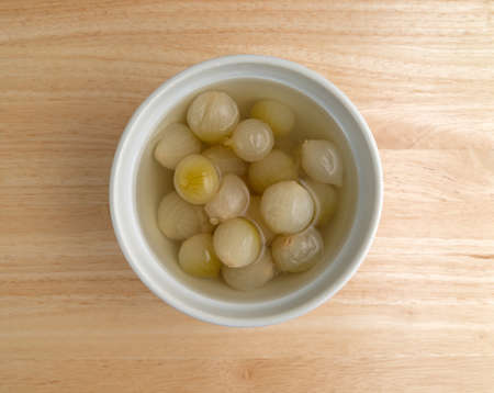 Top view of a serving bowl filled with cooked small onions in liquid on a wood table top illuminated with natural light.の写真素材