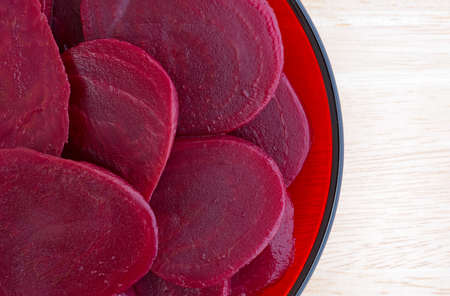 Top close view of sliced canned beets on a red plate atop a wood table top illuminated with natural...
