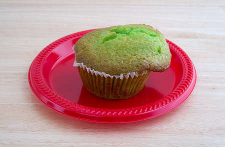 A freshly baked pistachio breakfast muffin on a red plastic plate atop a wood tabletop illuminated with natural light.の写真素材