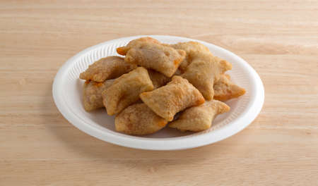 A serving of bite size pizza rolls on a white foam plate atop a wood table top illuminated with...