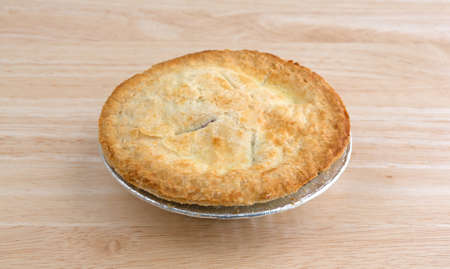 A small personal size fruit filled pie in a tinfoil dish on a wood table top illuminated with...