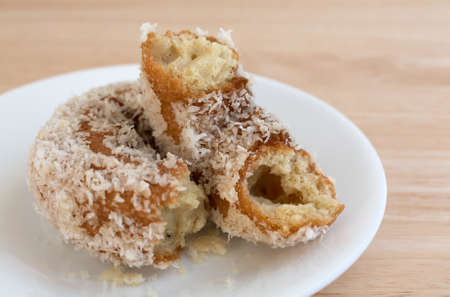 A coconut flaked plain donut that has been broken in half on a white plate atop a wood table top...