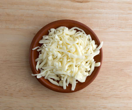 Top view of a portion of low moisture whole milk mozzarella pizza cheese in a small bowl atop a wood...