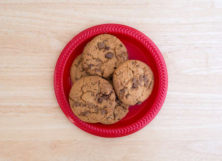 Top view of milk chocolate chip cookies on a plastic red plate atop a wood table top illuminated...