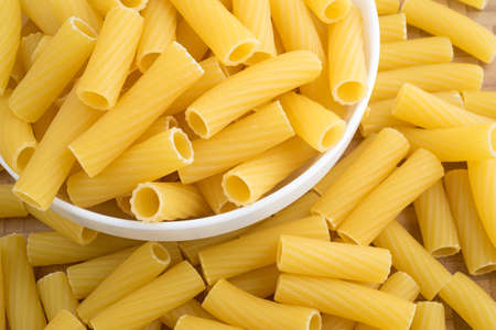 Top close view of a small white bowl filled with rigatoni pasta with more pasta strewn atop a wood...