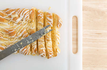 Top view a large cheese danish on a plastic cutting board with a slicing knife atop a wood table...