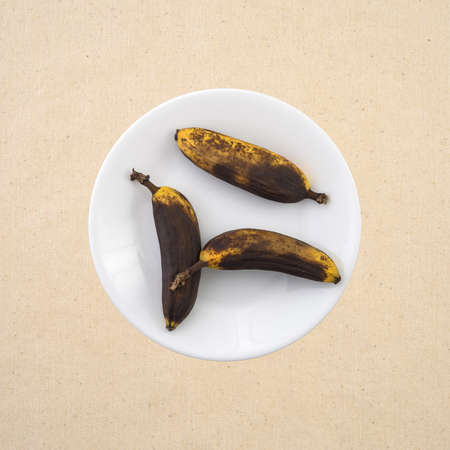 Top view of three small ripe bananas isolated on a white plate atop a beige tablecloth.の写真素材