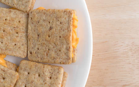 Top close view of several organic whole wheat crackers with canned cheese on a white plate atop a wood table.の写真素材