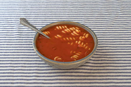 Rotini tomato soup in an old stoneware bowl with a spoon inserted into the food on a blue striped tablecloth.の写真素材