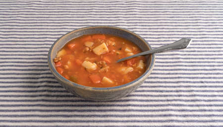 A serving of Manhattan style clam chowder in an old stoneware bowl with a spoon inserted into the food on striped tablecloth.の写真素材