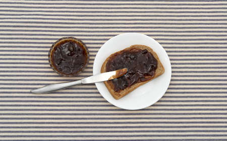 Top view of plum preserves spread on a piece of wheat bread atop a white plate with a butter knife...
