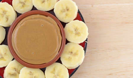 Top close view of sliced bananas on a red plate in a circle around a bowl of peanut butter on a wood table.の写真素材