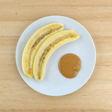 Top view of a sliced banana with a portion of creamy peanut butter on a plate atop a wood table.の写真素材