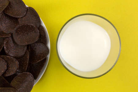 Top close view of a plate of dark chocolate covered rice crisp wafers with a glass of milk to the side atop a yellow tablecloth.の写真素材