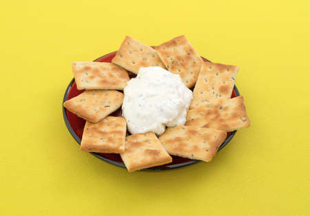 Rosemary and olive oil seasoned crackers with French onion dip on a red plate atop a bright yellow...