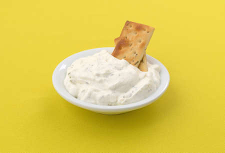 Rosemary and olive oil seasoned crackers inserted into French onion dip in a small bowl on a bright yellow tablecloth.の写真素材