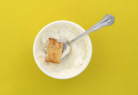 Top view of a container of French onion dip with a cracker atop a spoon in the food atop a bright yellow tablecloth.の写真素材