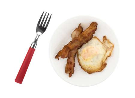 Top view of a fried egg with two strips of bacon on a plate with a fork to the side isolated on a white background.の写真素材