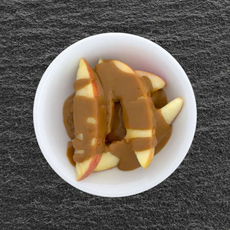 Slices of red apples covered with creamy peanut butter in a small bowl atop a black slate table top.の写真素材