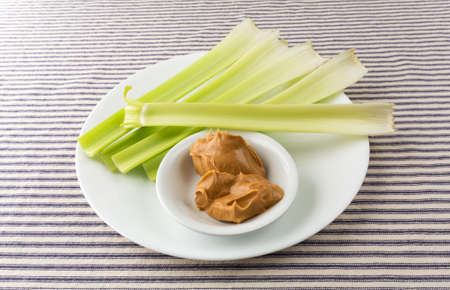 Several celery stalks with a bowl of peanut butter on a white plate atop a blue striped tablecloth.の写真素材