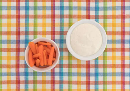 Top view of several small peeled carrots in a bowl with ranch dressing to the side atop a colorful...