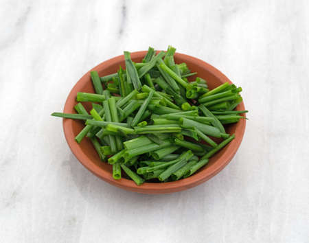 Cut organic chives in a small red clay bowl atop a gray marble cutting board.