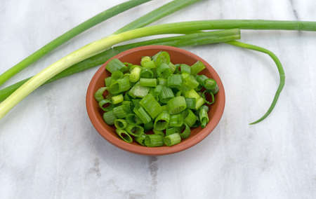 A small red clay bowl filled with freshly chopped green onions plus several leaves on a gray marble counter top.の写真素材
