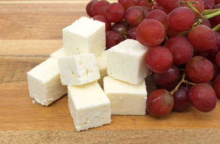 Several pieces of feta cheese on a wood cheese board with a bunch of fresh red grapes.