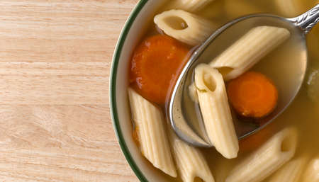Top view of a small bowl filled with penne pasta, carrots and celery soup in a chicken broth plus a spoon in the food atop a brown wood table.の写真素材