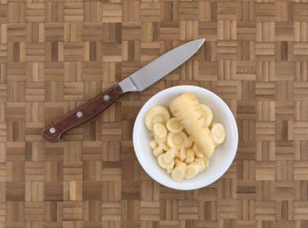 Top view of a small filled with parsnip slices plus a knife to the side on a wood cutting board.