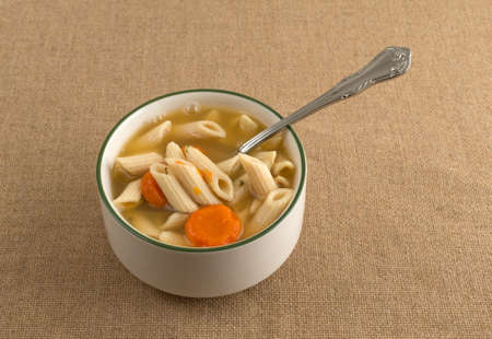 A small bowl filled with penne pasta, carrots and celery soup in a chicken broth plus a spoon in the food on a brown burlap tablecloth.の写真素材
