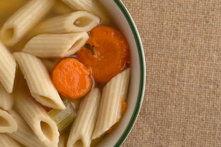 Top close view of a small bowl filled with penne pasta, carrots and celery soup in a chicken broth on a brown burlap tablecloth.の写真素材