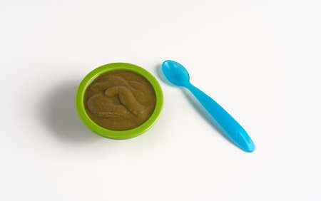 Top view of a serving of spinach baby food in a green bowl with a spoon to the side on a white table top illuminated with window light.の写真素材