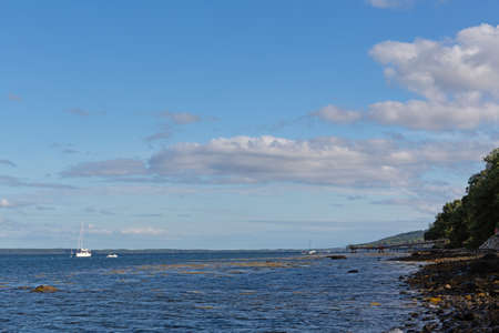 The rocky shoreline of Belfast, Maine on a cloudy summer day with distant boats at their moorings...
