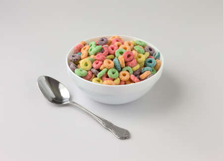 A white bowl filled with generic dry sugar coated fruity flavored breakfast cereal with a spoon to...