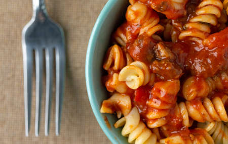 Top close view of a bowl of rotini pasta in a sausage and tomato sauce with a fork to the side on a brown tablecloth.の写真素材