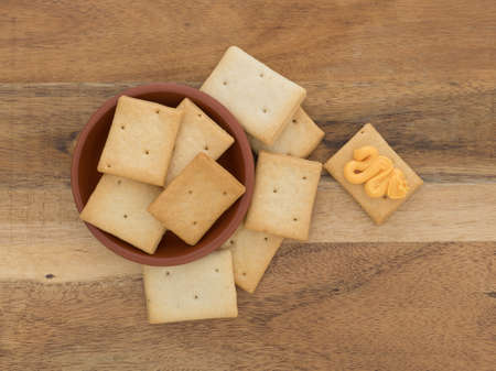 Top view of a serving of hard bread crackers in a red clay bowl with one to the side having canned cheese on a wood table.の写真素材