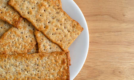 Top close view of multigrain flatbread crackers on a white plate atop a wood cutting board.