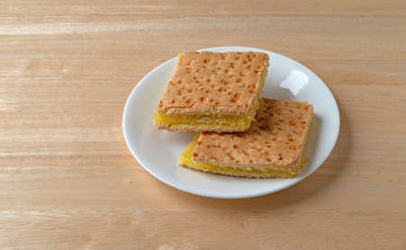 Two freshly baked lemon squares on a white plate atop a wood table illuminated with window light.