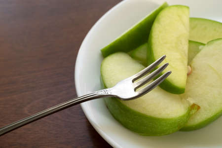 Close view of green sliced apples on a white plate atop a brown wood table with a fork on the fruit.の写真素材