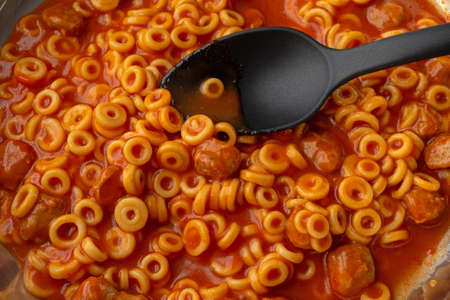 Canned spaghetti rings with meatballs cooking in a pan with a single ring on a black spoon.