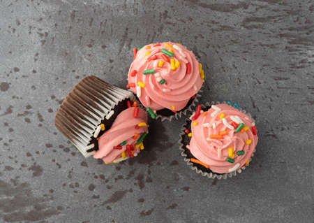 Top view of three pink frosted chocolate cupcakes with one tipped over on a gray background.