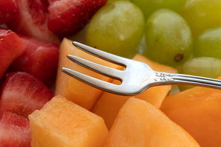 Close view of strawberries grapes and cut cantaloupe on a plate with a small fruit fork atop the food.の写真素材
